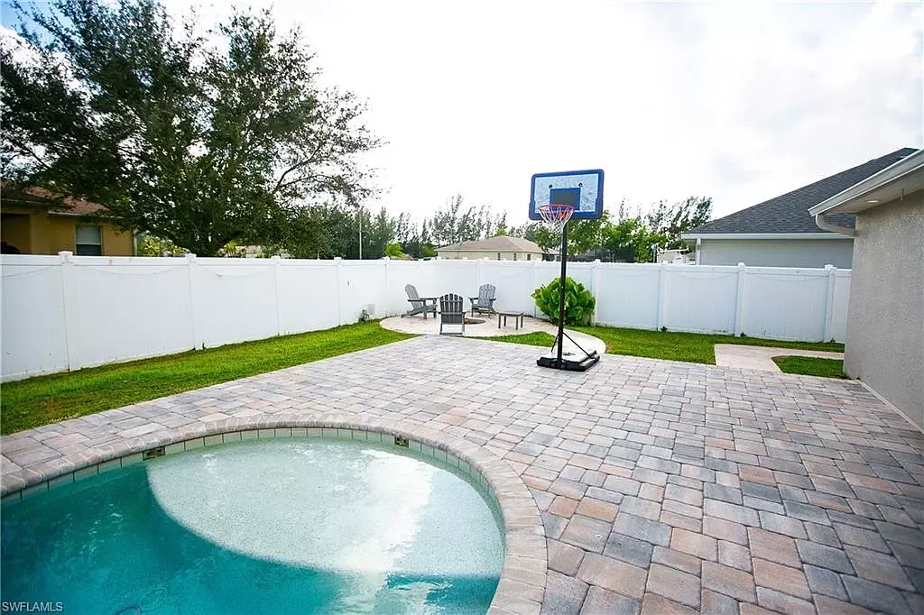 Backyard patio with pool, basketball hoop, chairs, and white fence.