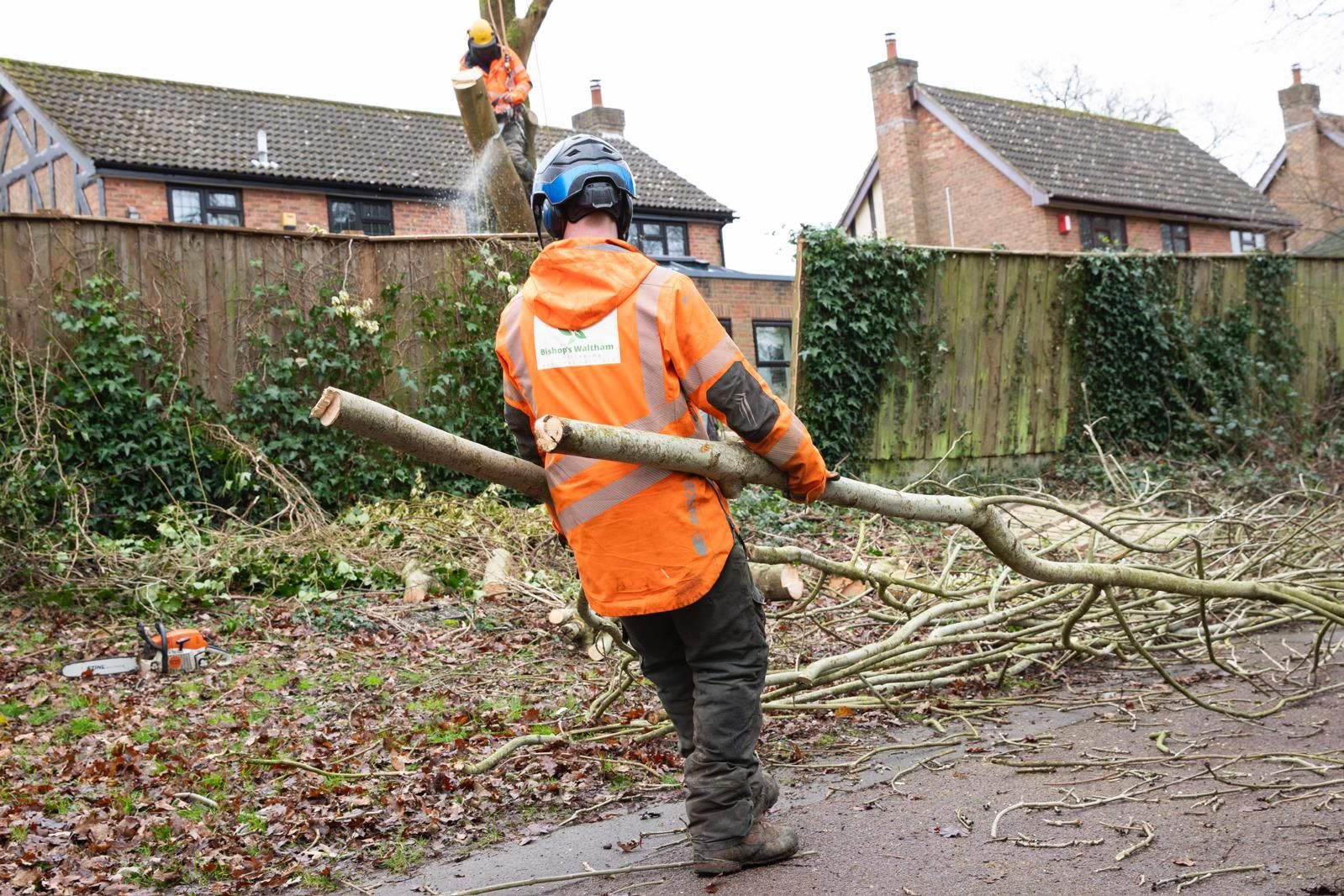 A tree surgeon in an orange jacket is carrying a large log.