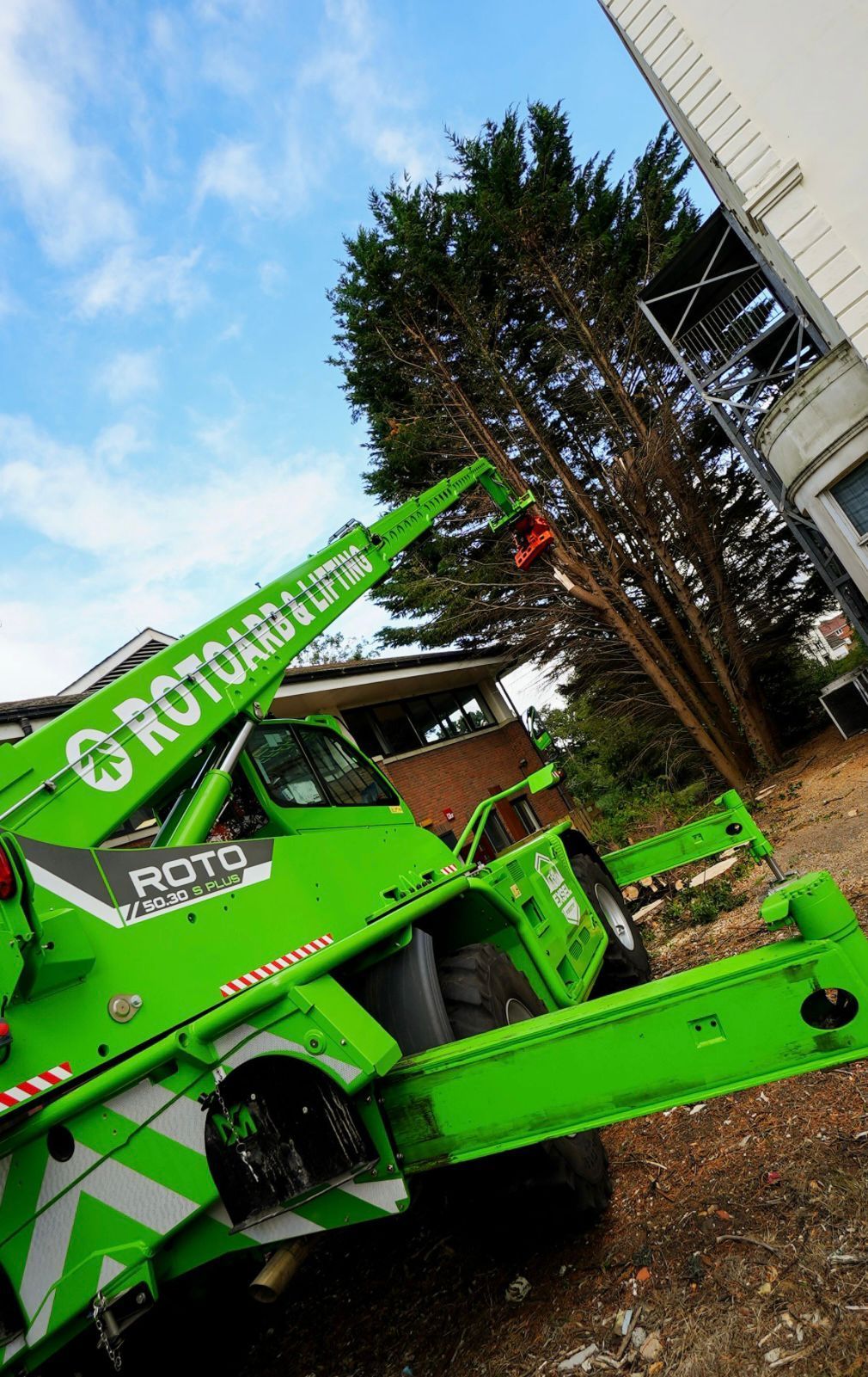Bright green ROTRANCH crane lifting near a building and tall tree outdoors
