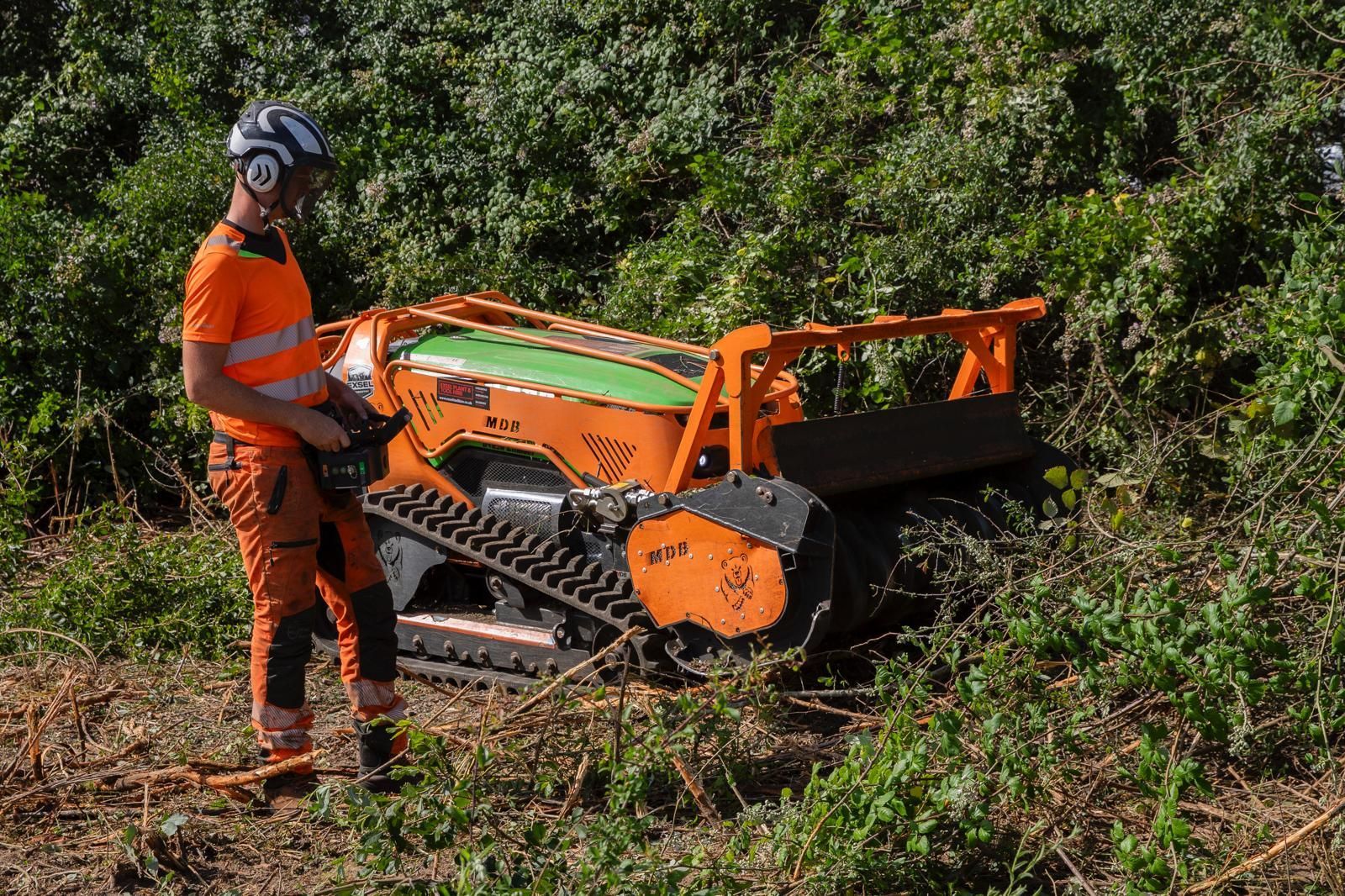 Worker in orange safety gear operating a green and orange tracked forestry machine on a hillside.