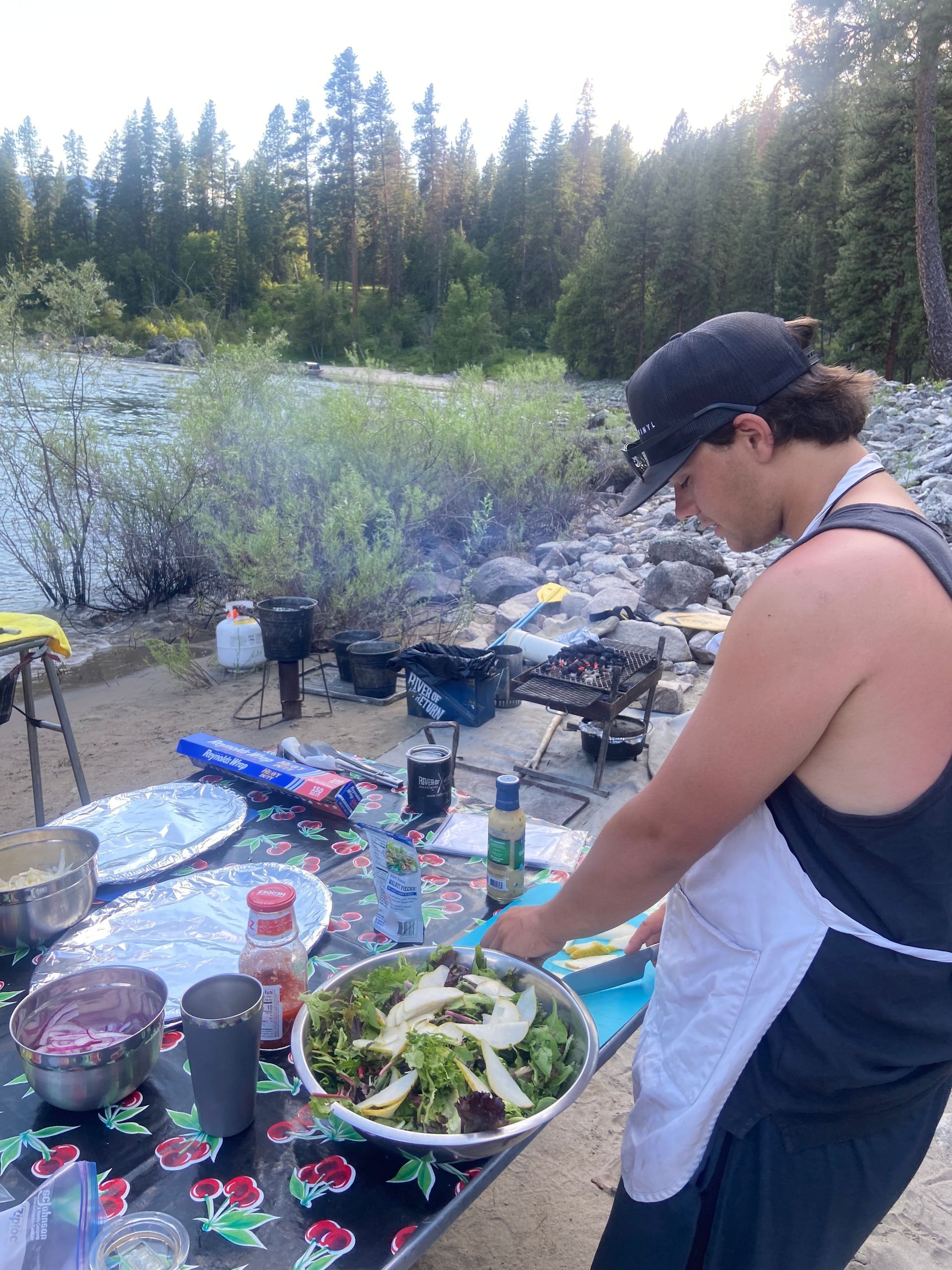 A man is preparing a salad on a table with a river in the background.