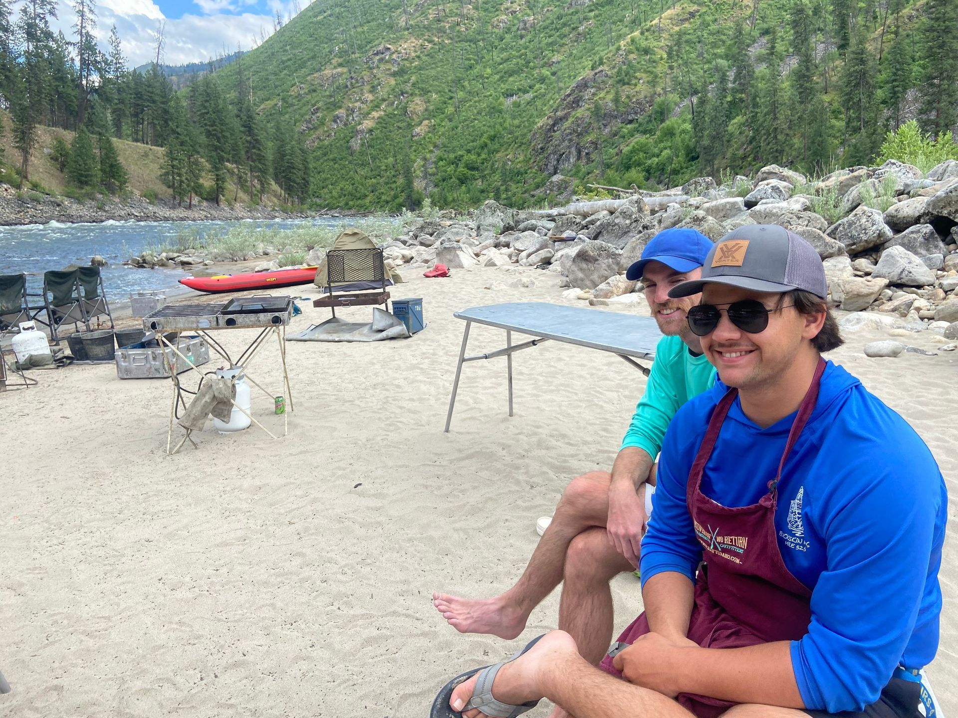 Two men are sitting on a sandy beach near a river.