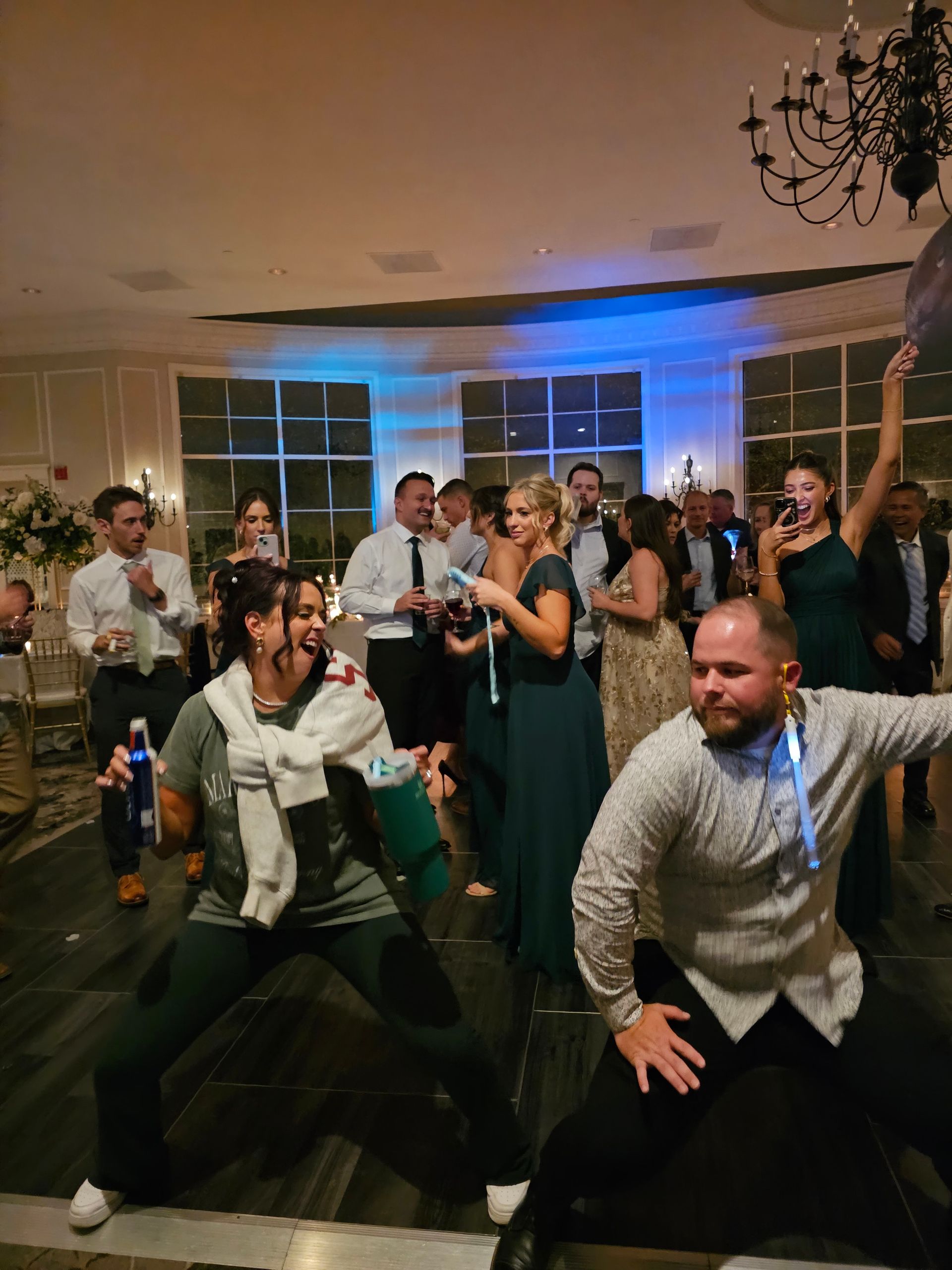 People dancing at a wedding reception. Some are holding drinks, illuminated lights, and balloons. The room has large windows.
