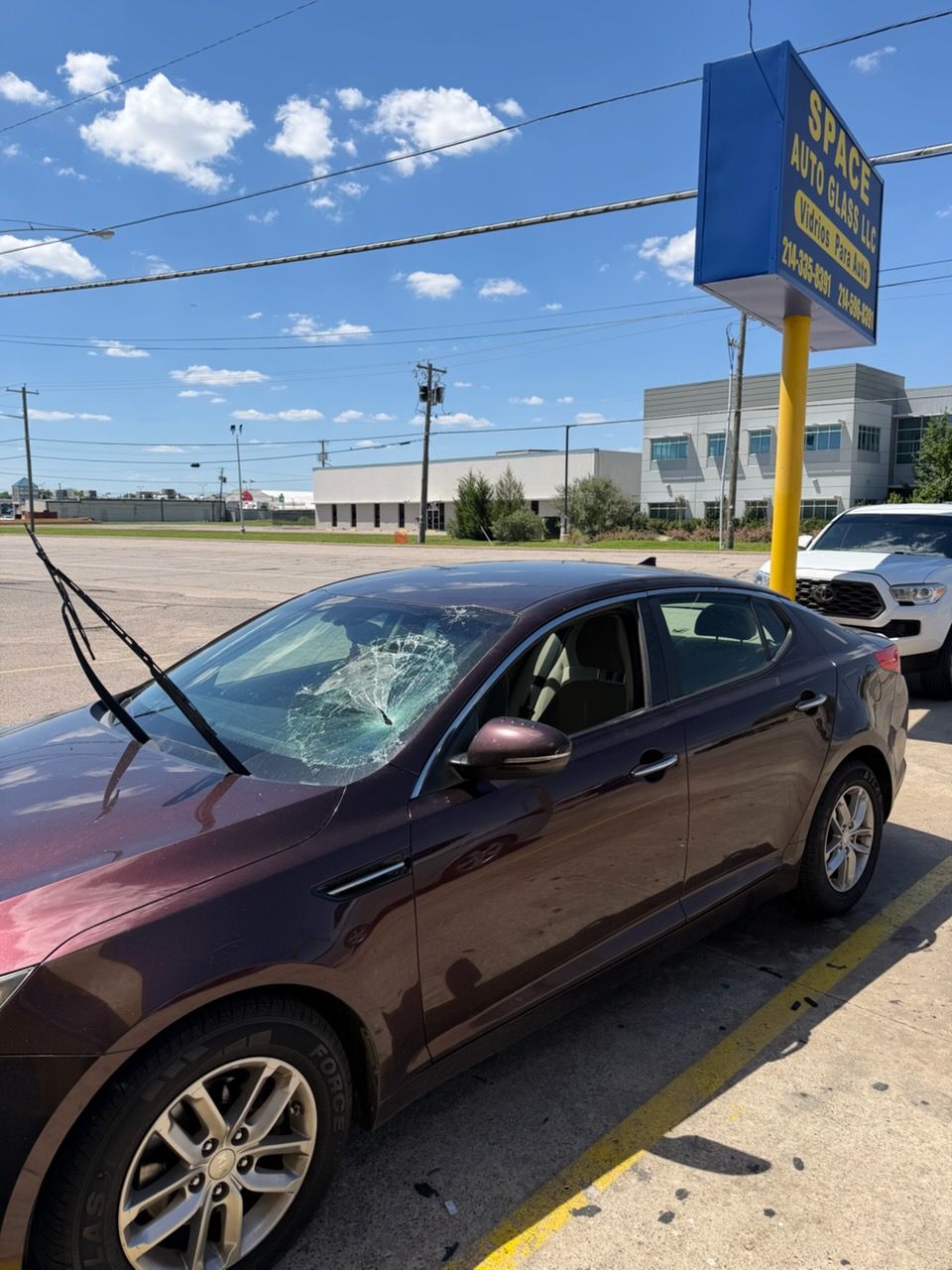 Maroon sedan parked by a curb with windshield wipers raised on a sunny street near a blue sign.