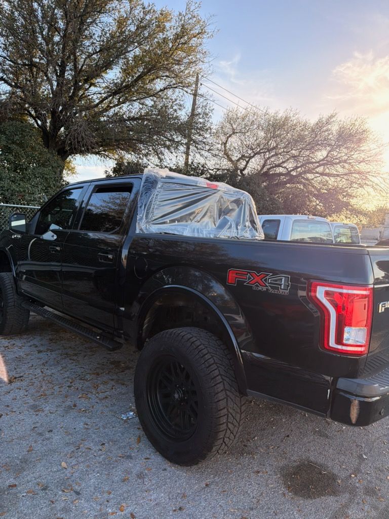 Black lifted pickup truck parked outdoors with rear window covered in plastic wrap near trees at sunset