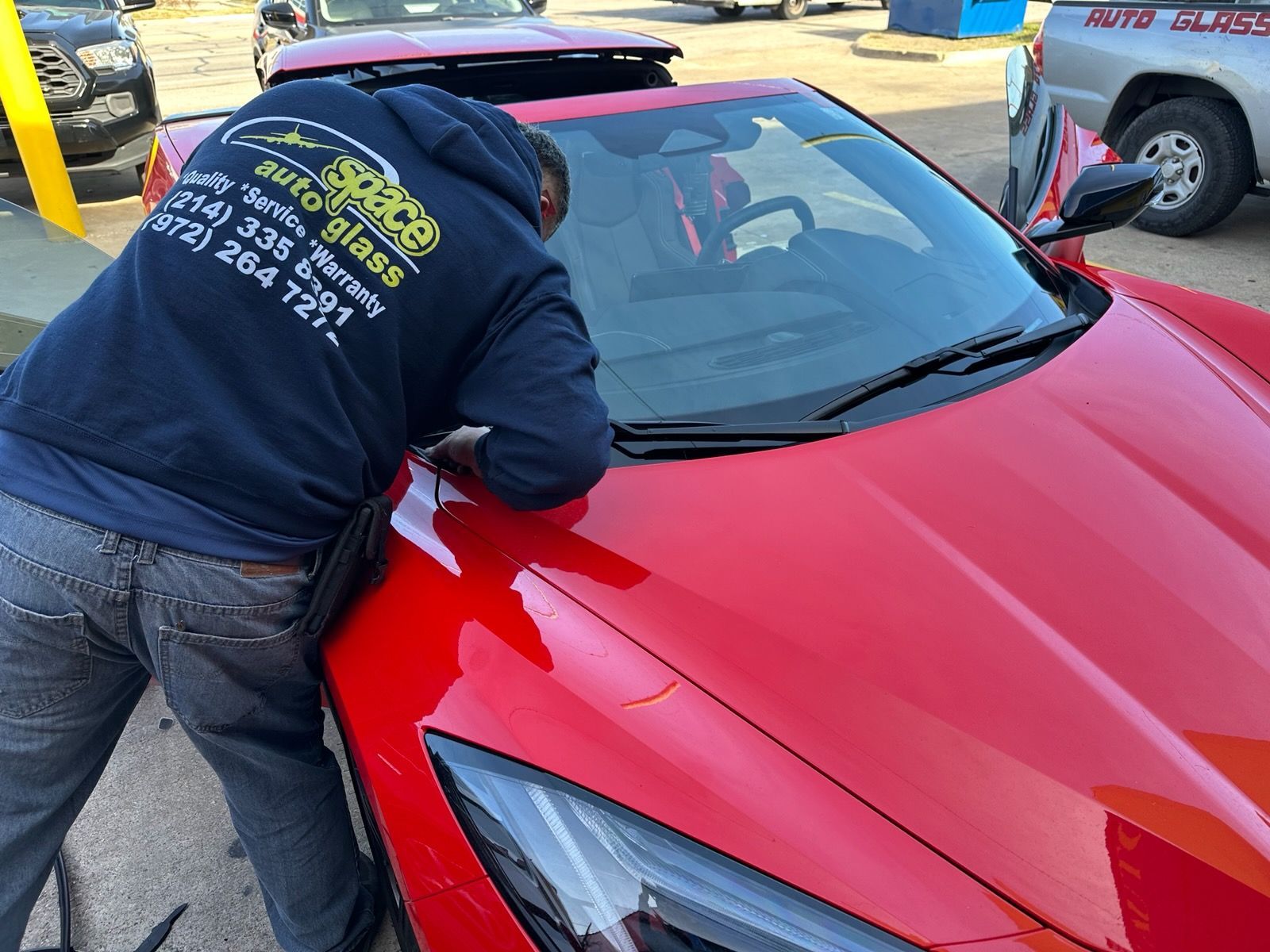 Person in a navy hoodie leaning over a red sports car, cleaning or inspecting the windshield.