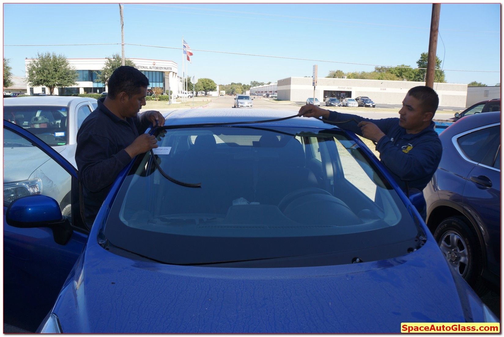 Two men cleaning a blue car windshield in a parking lot