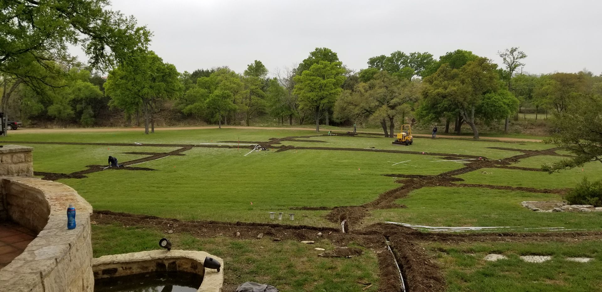 Park lawn with muddy runoff channels, trees, and a stone wall in the foreground