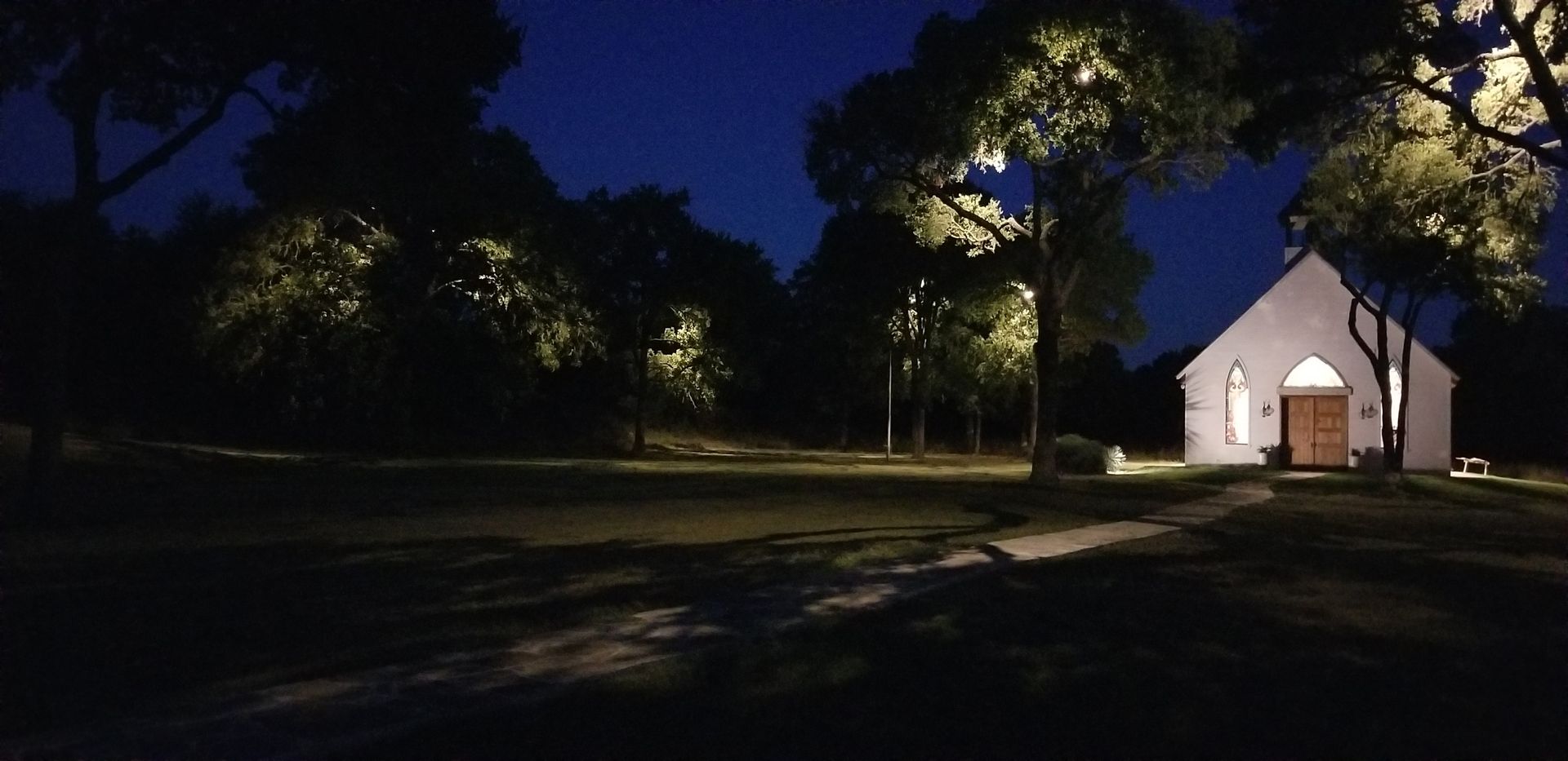 Small white chapel at night, lit by warm lights among dark trees and an empty parking lot