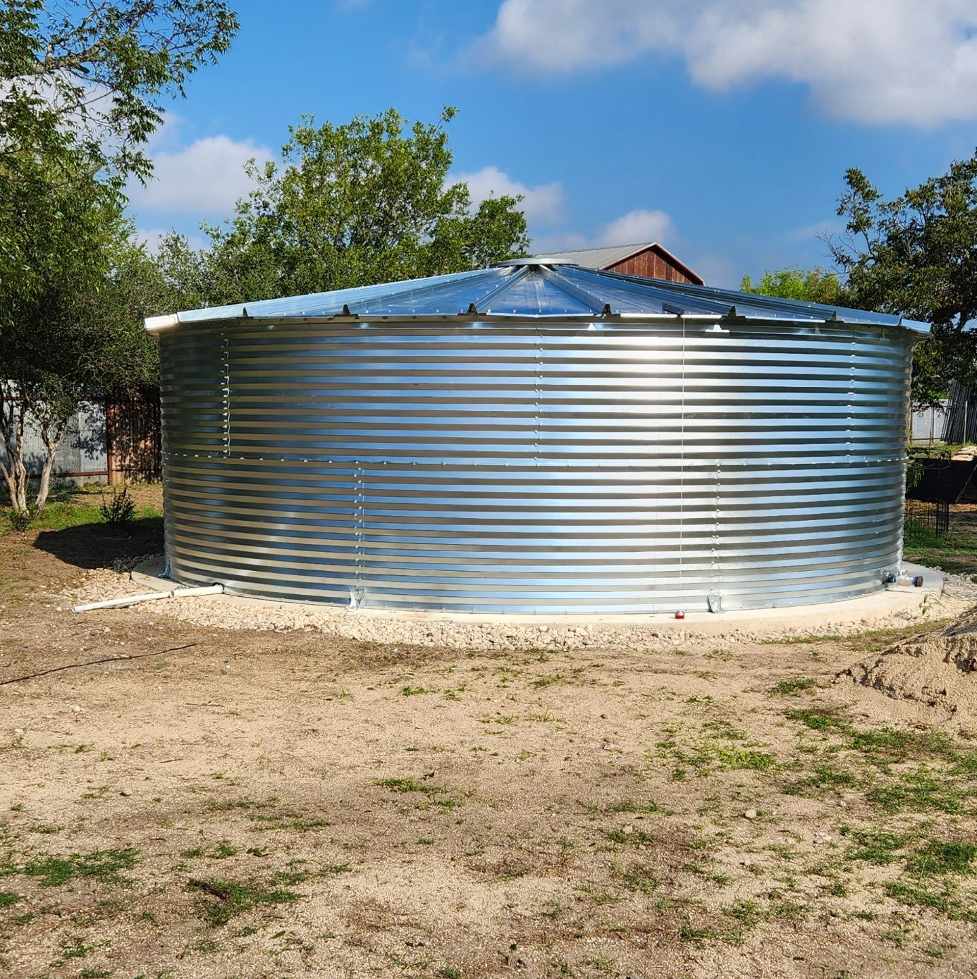 Large silver corrugated metal water tank in a grassy yard under a blue sky