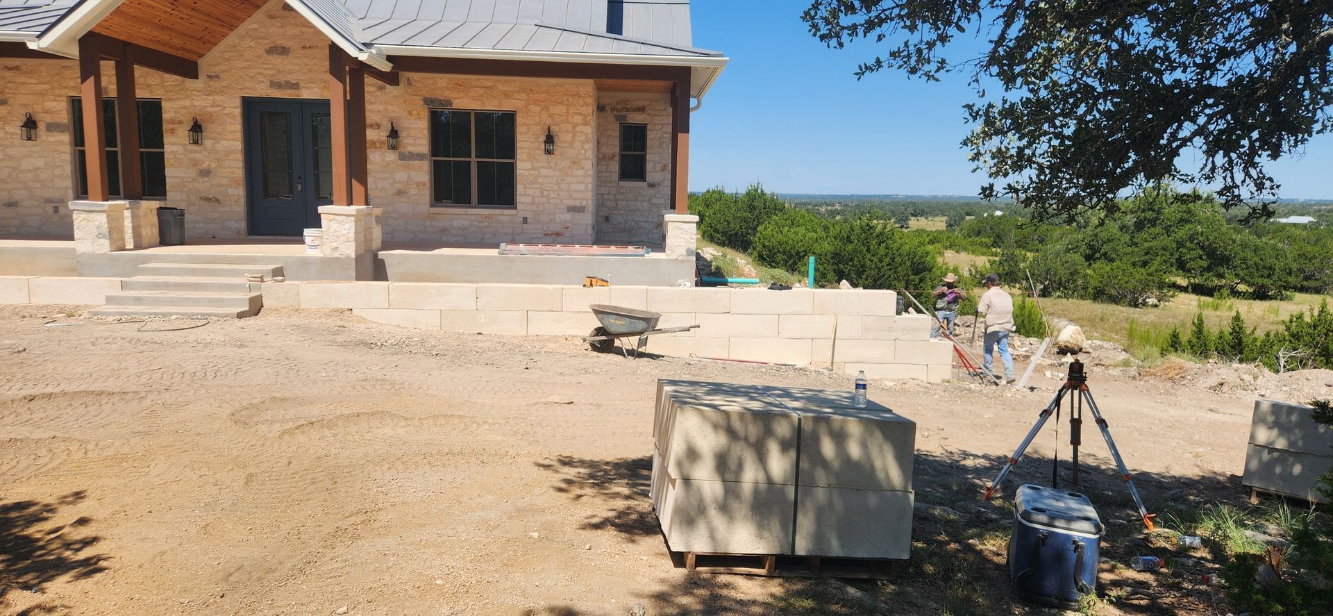 Stone house under construction with steps, dusty yard, and a tripod setup in the foreground