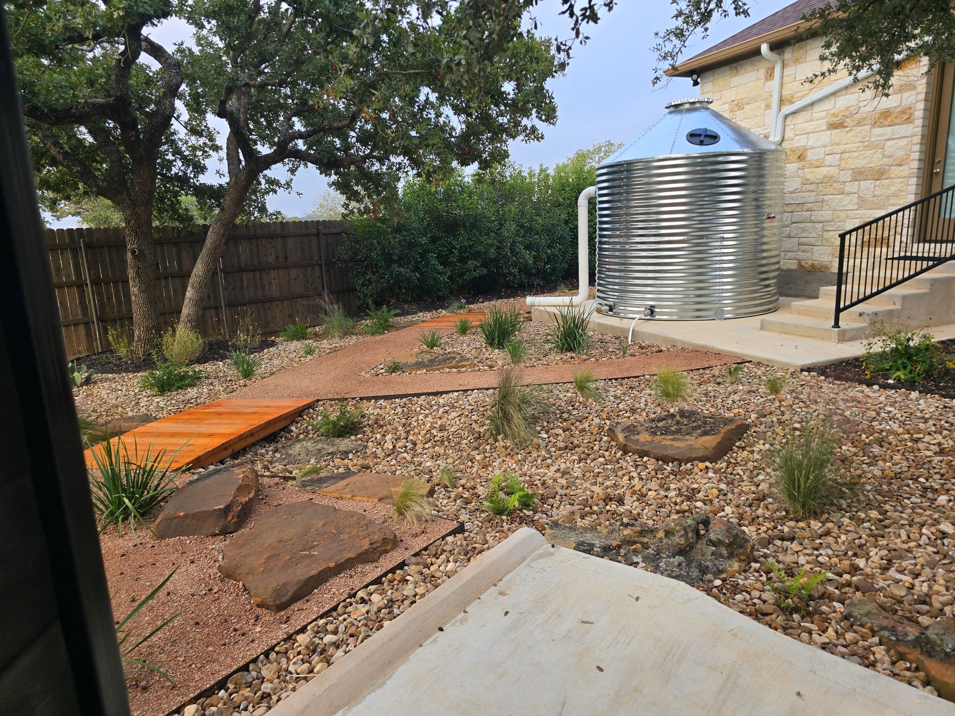 Rocky backyard garden with dry plants, a wooden fence, and a metal rainwater tank beside a stone house.