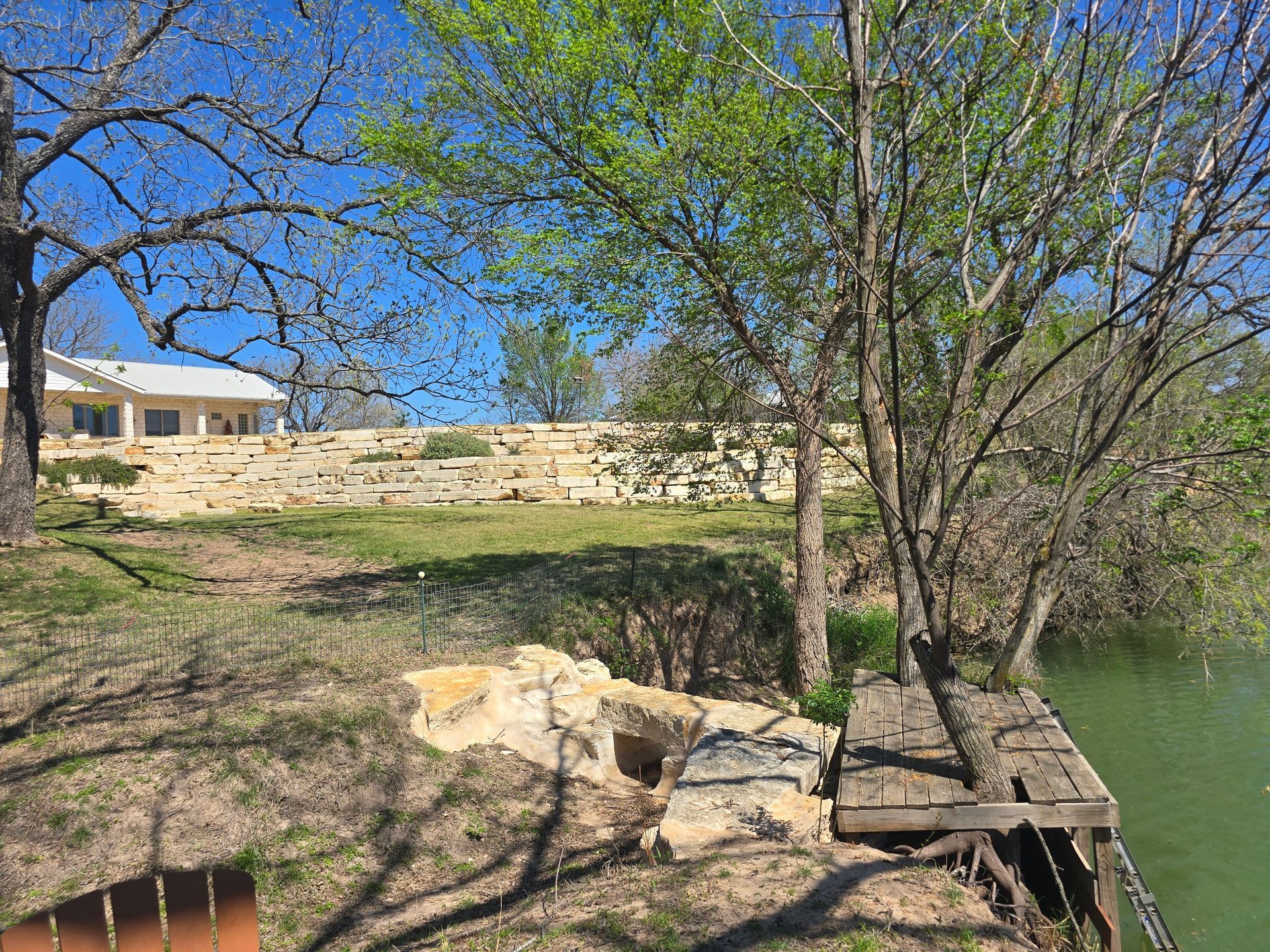 Rocky lakeshore with trees, a wooden dock, and a stone wall under a clear blue sky