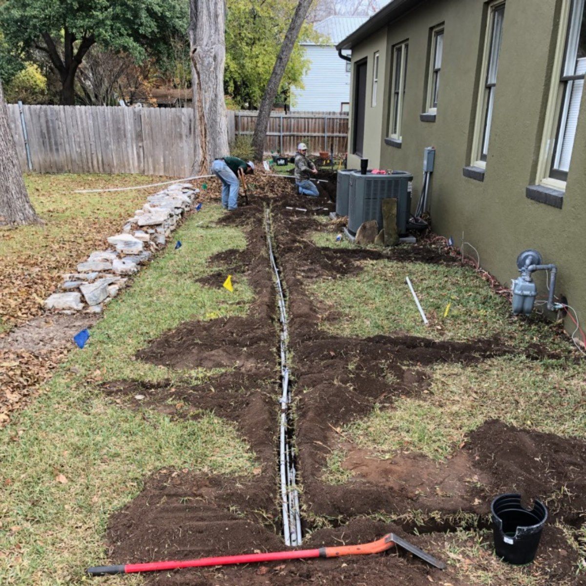 Backyard trench beside house with exposed utility line and a worker digging along the foundation