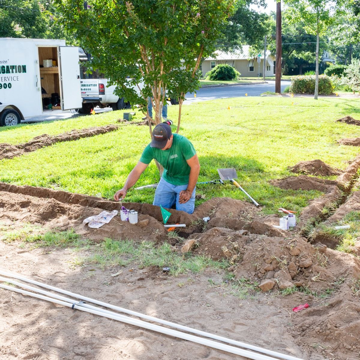 Worker shoveling dirt beside a trench in a grassy yard, with pipes and a truck nearby.