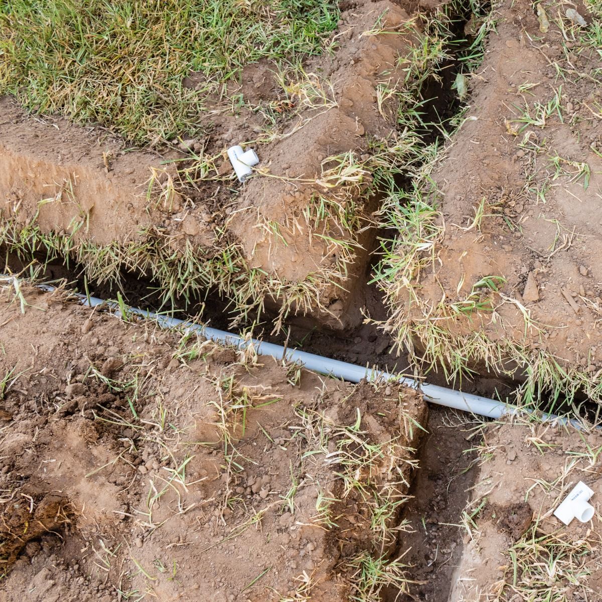 Broken gray pipe exposed in a dirt trench beside grass and a white fitting