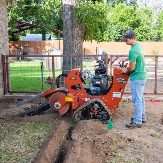Worker operating orange trencher cutting a narrow trench beside a yard fence