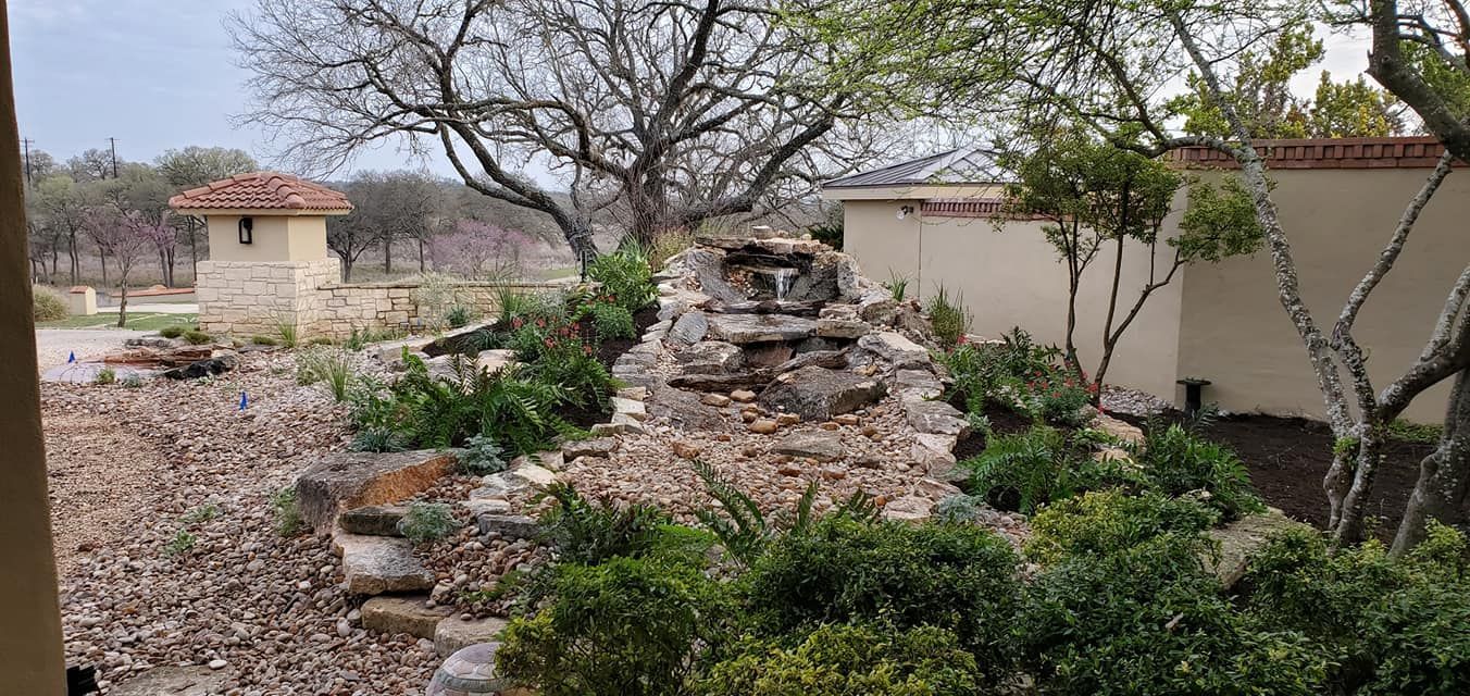 Desert landscaping with a rock waterfall feature, shrubs, trees, and tan buildings in the background