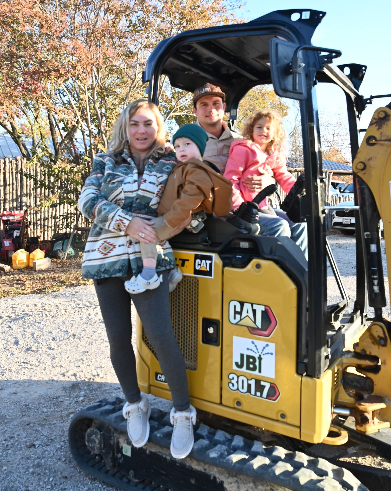 Family sitting on a CAT mini excavator outdoors in a gravel yard, with trees and pumpkins in the background