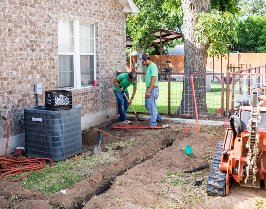 Workers digging a trench beside a brick house, with tools and an excavator in a yard under construction