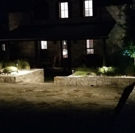 Nighttime view of a dark house with lit windows and a stone patio lined by low garden walls.