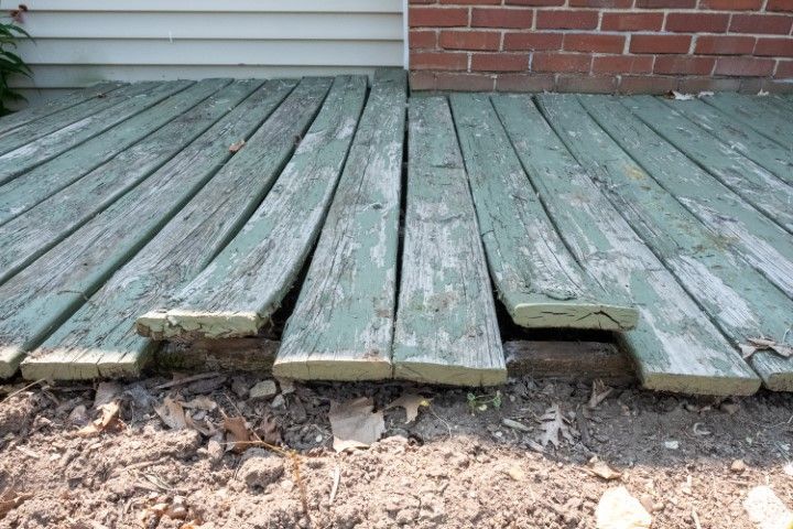 Weathered green wooden deck boards with loose, broken planks near a brick wall