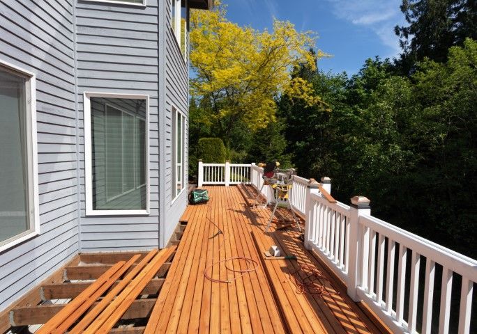 Wooden deck beside a gray house, with white railing and trees under a sunny sky.