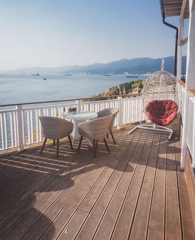 Sunny seaside deck with white chairs, a red heart cushion, and a view of the calm sea and distant mountains.