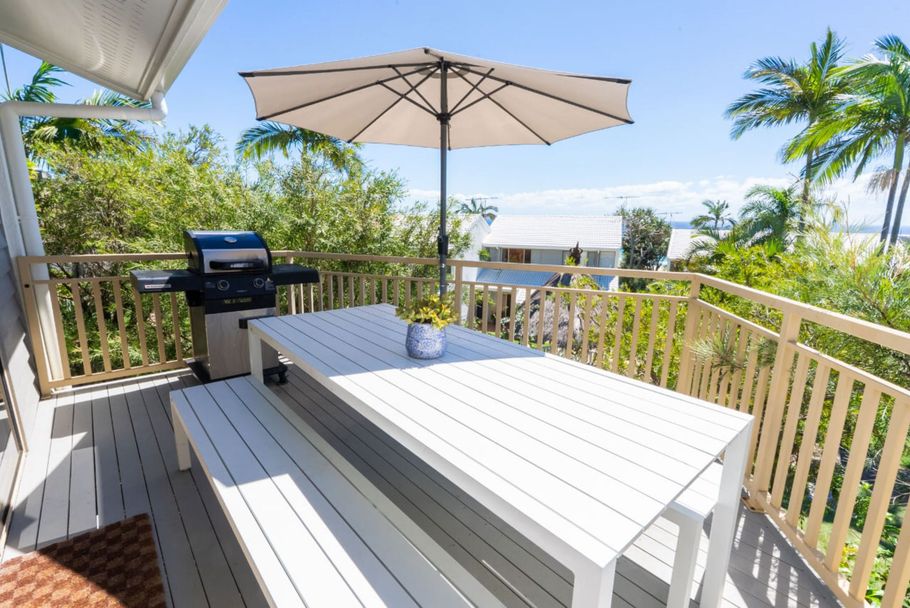 Sunny deck with white table, umbrella, grill, and tropical greenery overlooking the water