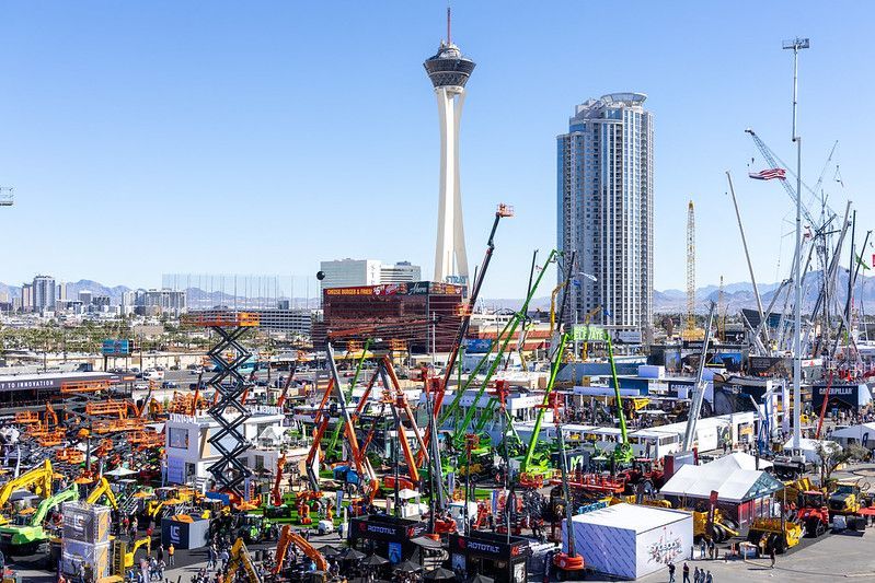 Crowded outdoor expo with colorful cranes and tents, city skyline, and tower in the background