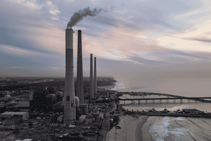 Industrial smokestacks beside a coastal power plant at dusk, with smoke rising over the shoreline and bridges.