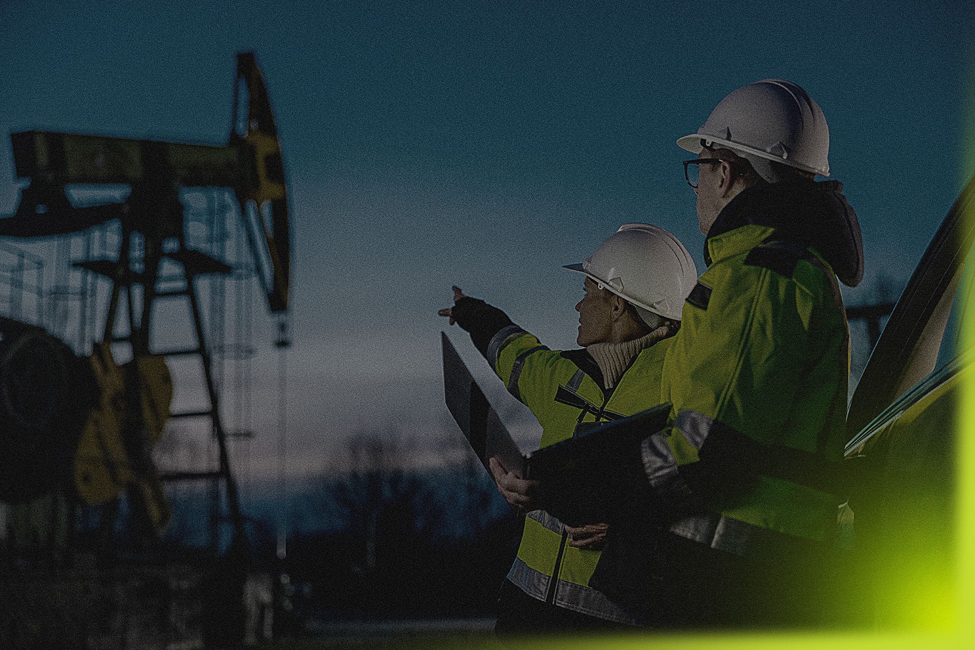Two engineers in hard hats and high-vis vests at an oil pump, one pointing at a diagram.