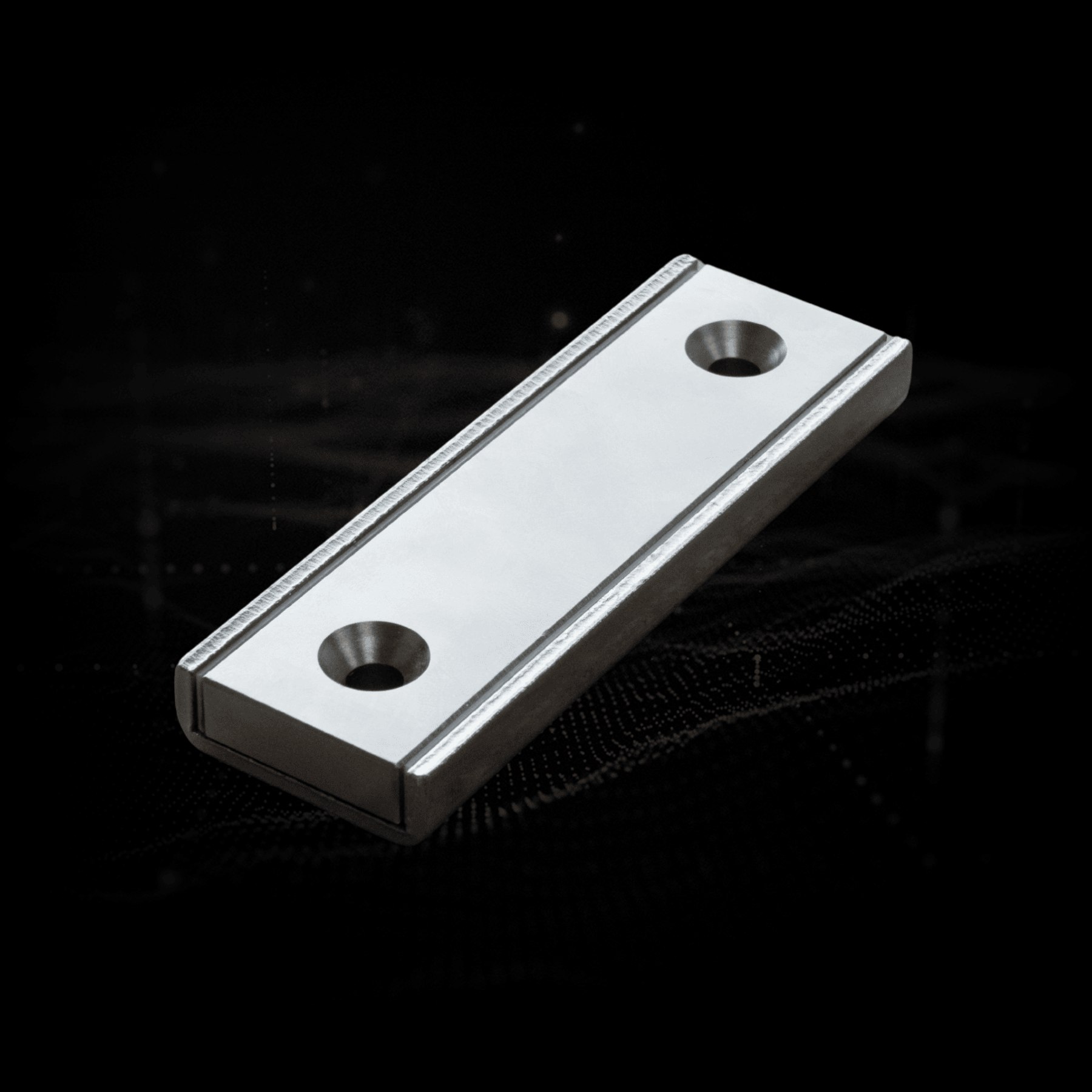 Silver rectangular magnet with two countersunk holes on a dark background