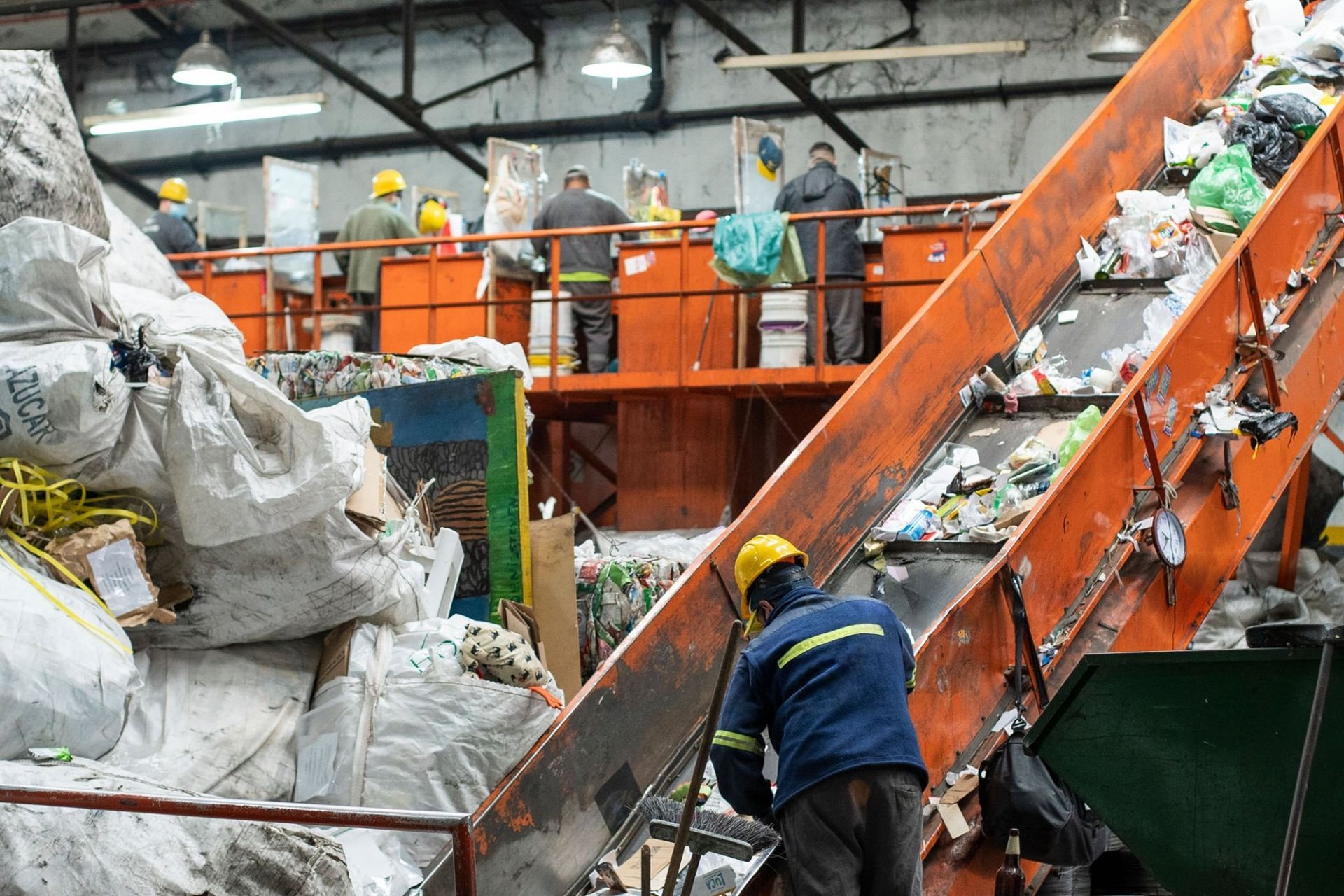 Workers sorting trash on a conveyor belt in a waste facility, with orange machinery and large garbage bags.