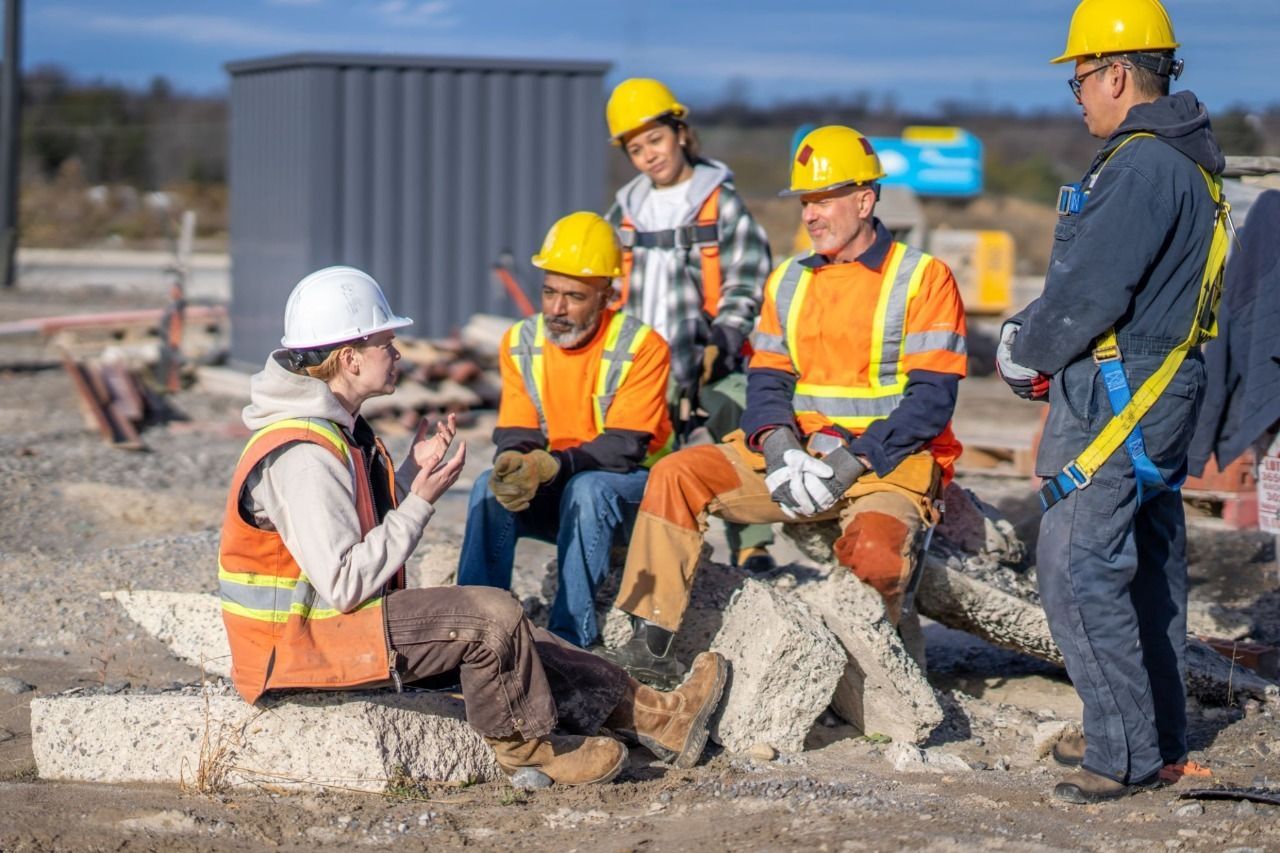 Five construction workers in high-visibility gear and hard hats sit and stand on a job site, having a group discussion.