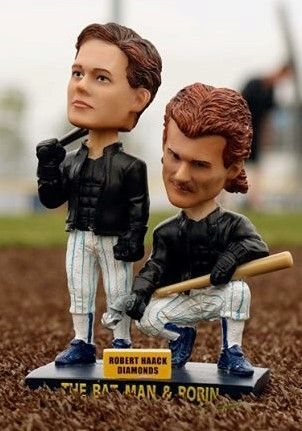Two bobblehead-style figures in black jackets and white pants, one holding a baseball bat on a display base.