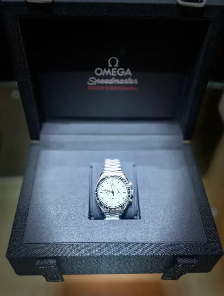 Omega Speedmaster watch in an open black presentation box on a dark background.