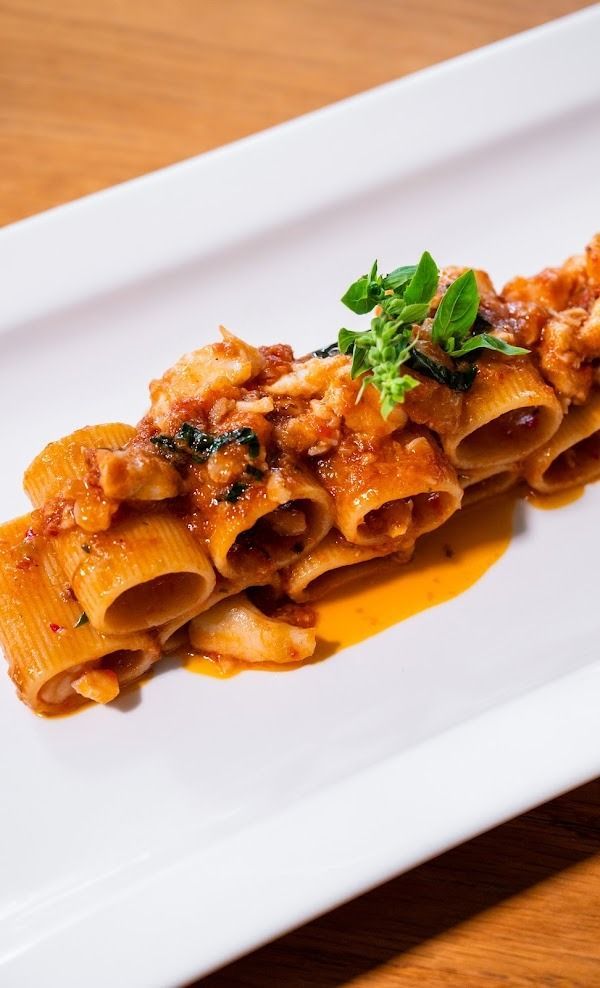 Pasta arrotolata in un ricco ragù di carne, guarnita con basilico e servita su un piatto rettangolare bianco.