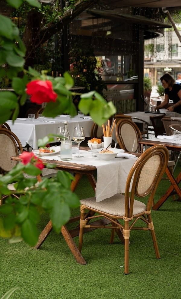 Tavolino da caffè all'aperto con tovaglia bianca, sedie in vimini e rose rosse su un patio verde.