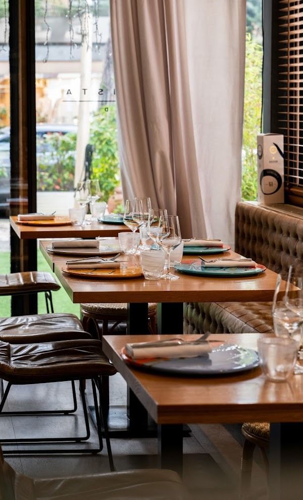 Tavoli vuoti in un ristorante, accanto a grandi finestre con tende beige e apparecchiati.