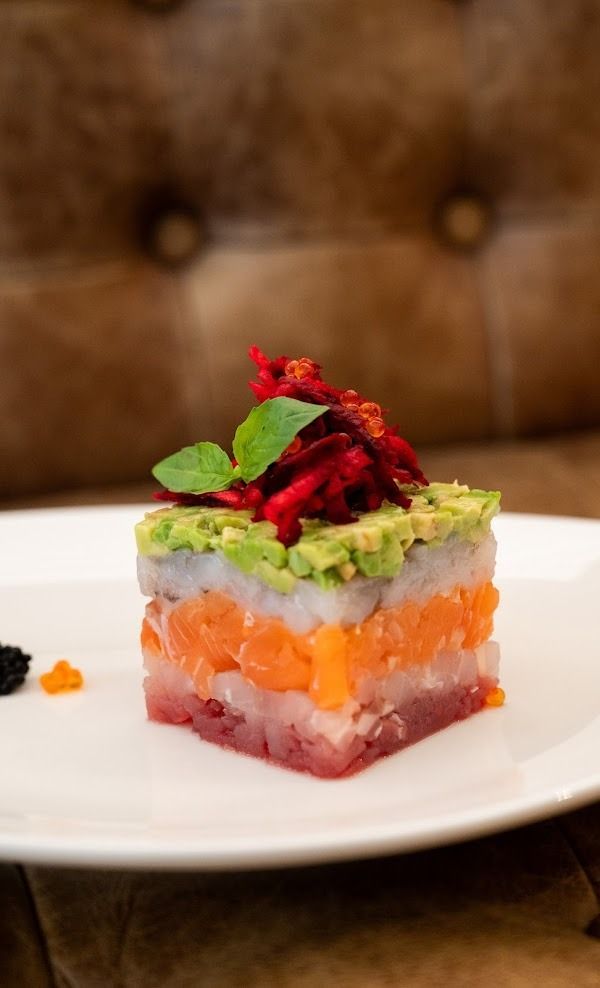 Dessert colorato a strati su un piatto bianco, guarnito con avocado e decorazione rossa, su uno sfondo marrone trapuntato.