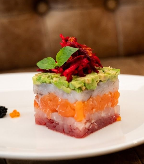Torretta di tartare di salmone a strati guarnita con avocado ed erbe aromatiche su un piatto bianco.