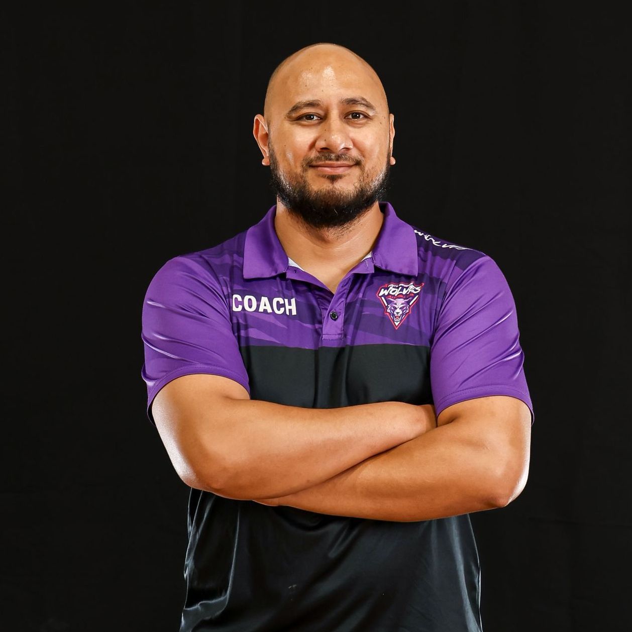 A man in a purple shirt and black shorts is standing with his arms crossed.