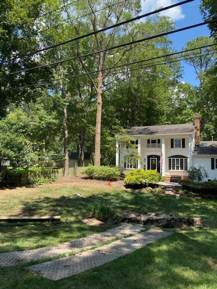 White house with front porch in a leafy yard, seen from a stone path under tall trees.