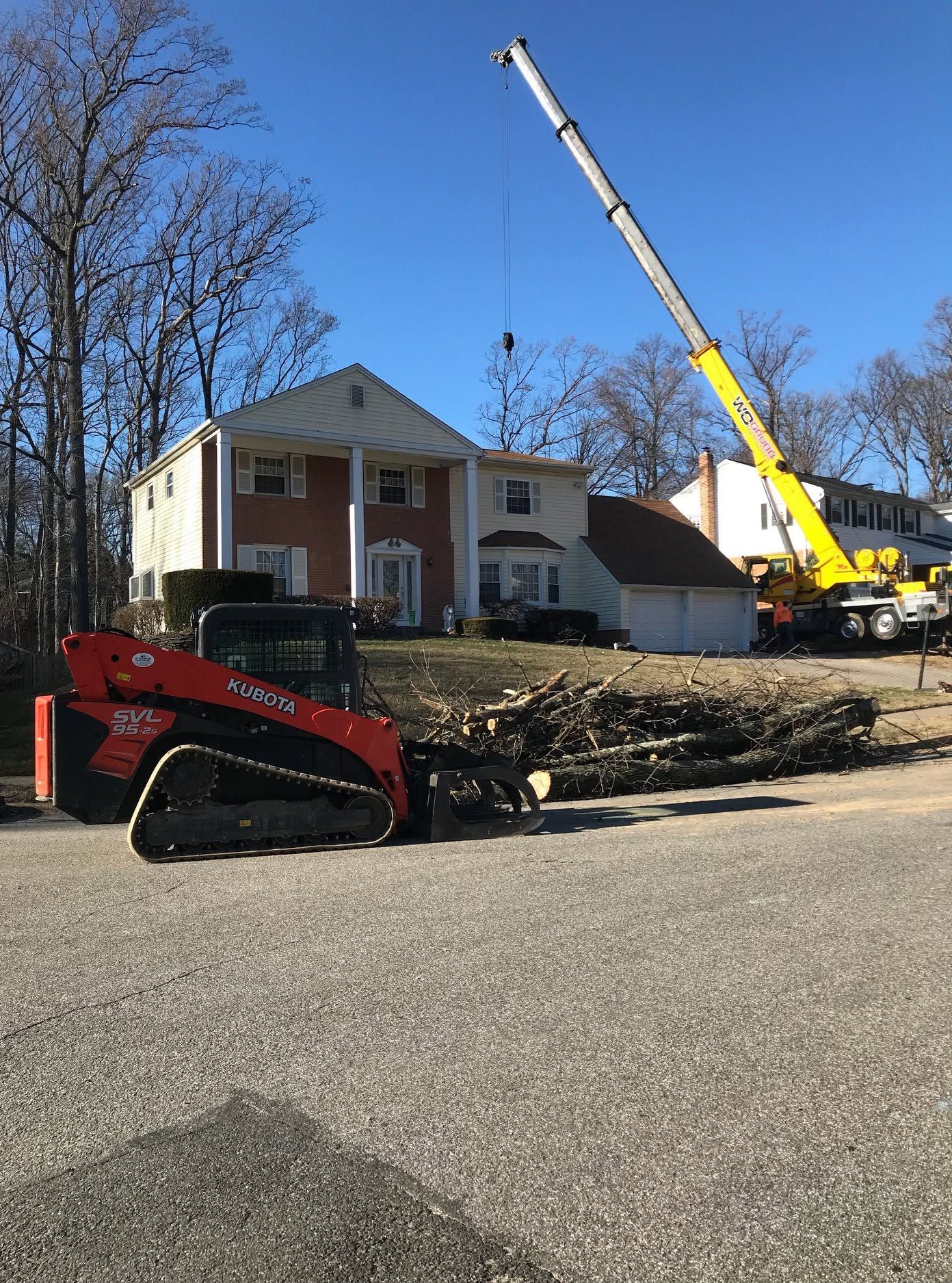 Red excavator in front of a brick house with a crane lifting a roof section on a clear day