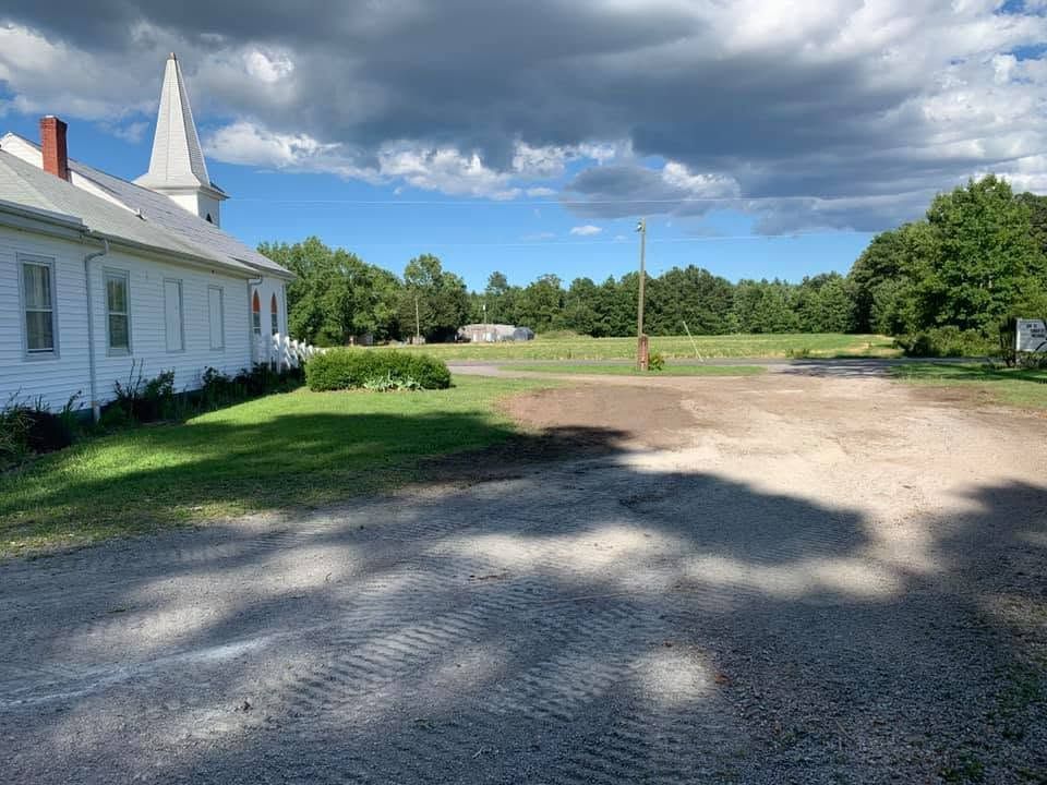 White church building beside a gravel lot under a cloudy sky, with green trees in the background