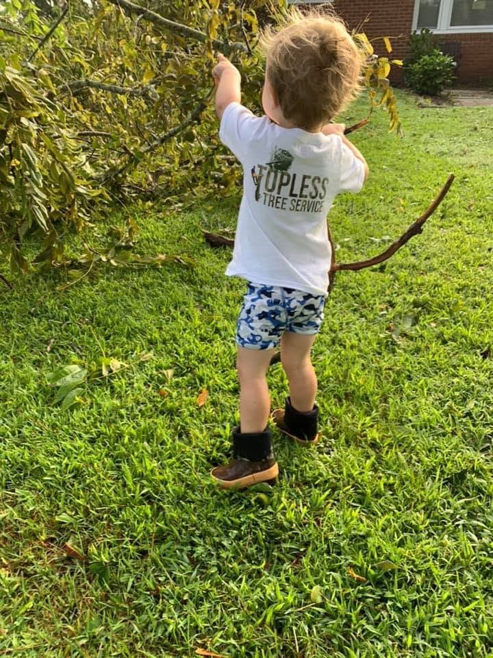 Child in a white shirt and blue shorts reaching up toward a tree in a green yard.