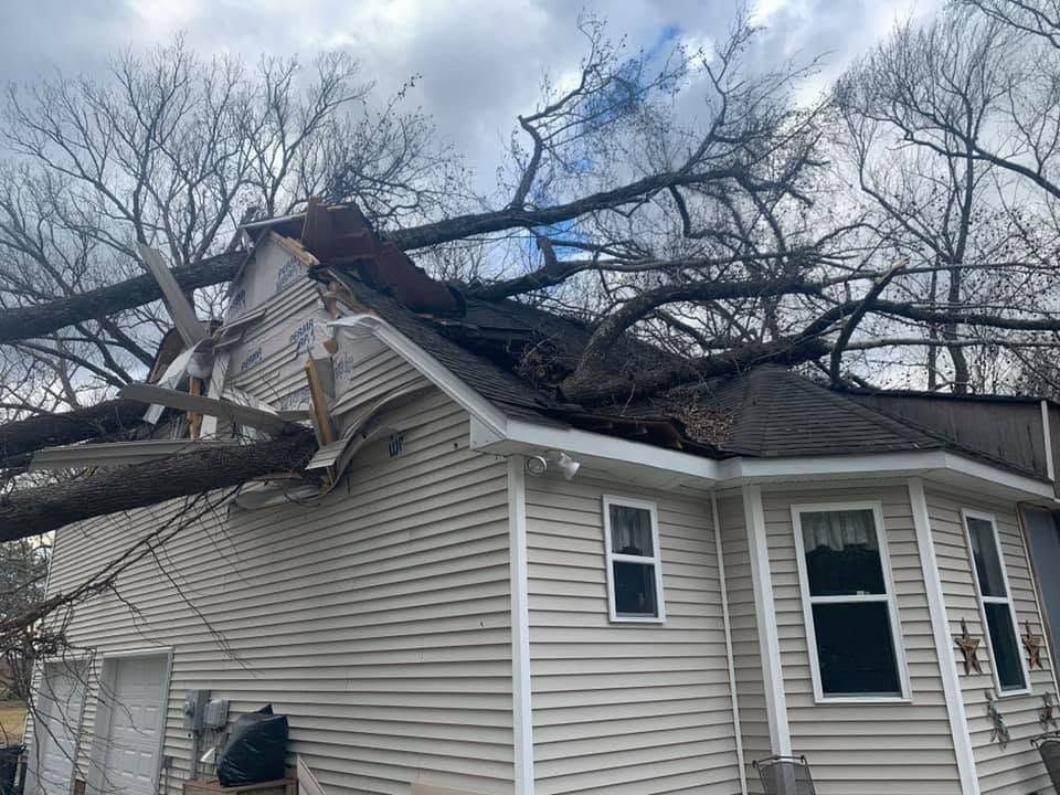 Tree fallen on house roof, damaging beige siding under cloudy sky