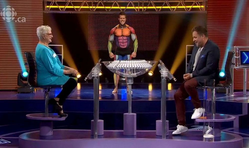 A man in a superhero costume is standing in front of a table with two people sitting around it.