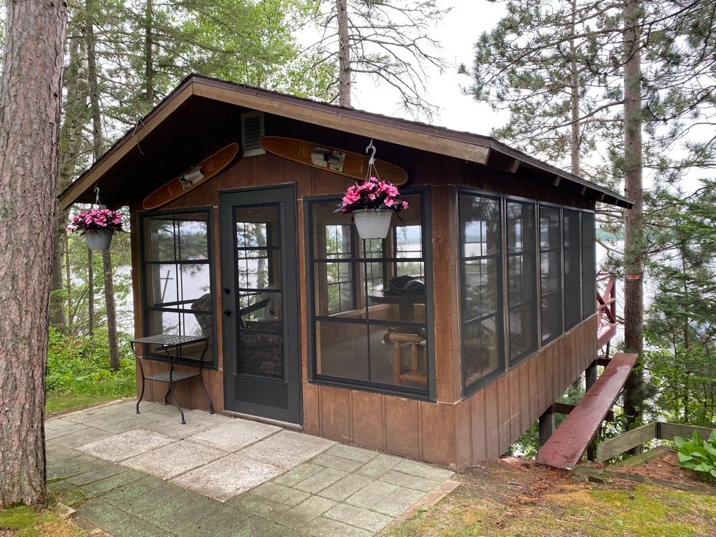 Small brown cabin with screened porch and hanging pink flower baskets in a wooded area